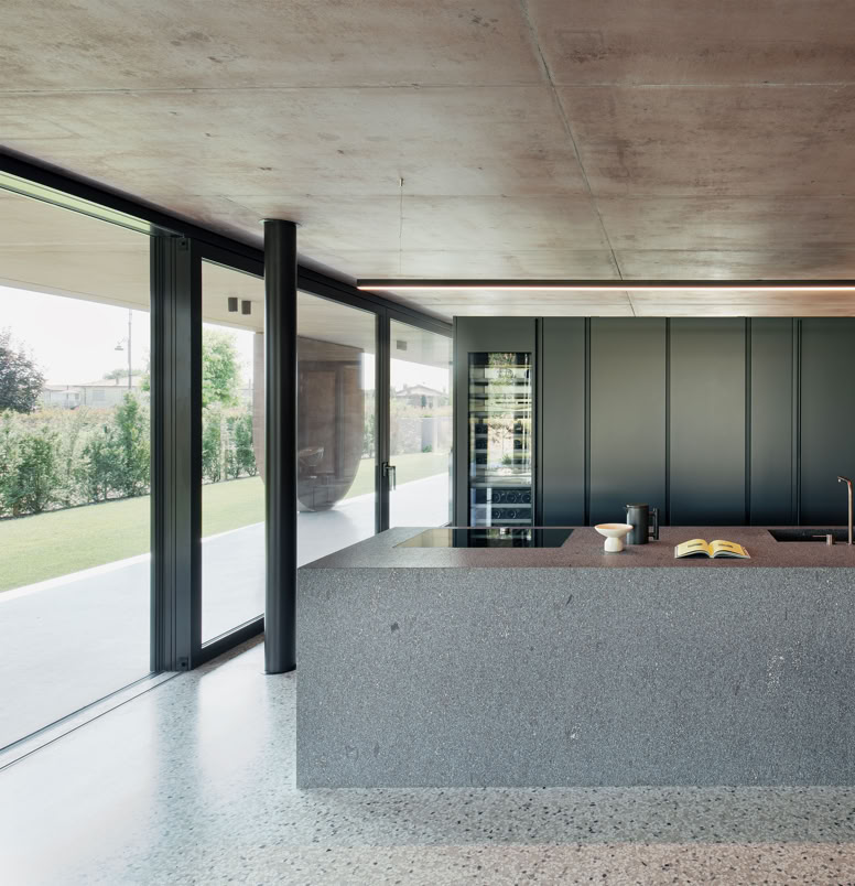 Open-plan kitchen of the villa designed by MIDE architetti, where colored concrete interacts with surrounding vegetation, creating a suspended atmosphere between interior and exterior.