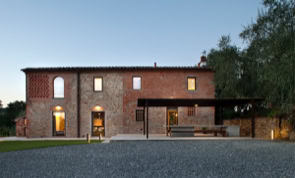 Vista serale della facciata in pietra e mattone faccia a vista, con infissi in acciaio e scuri in legno che valorizzano l’architettura tradizionale toscana.